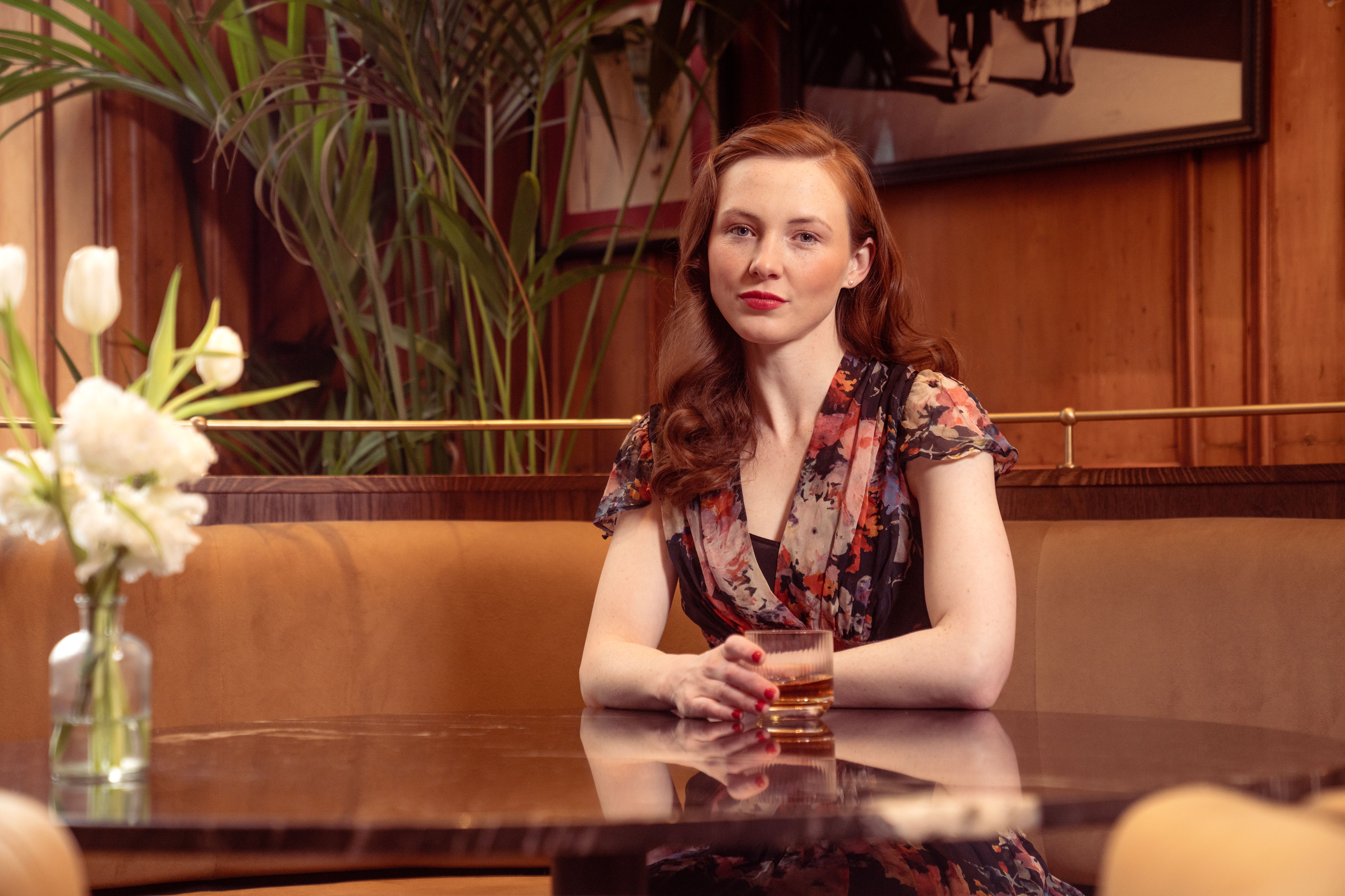A fashionable woman with red hair sips a drink, seated at a stylish restaurant table.