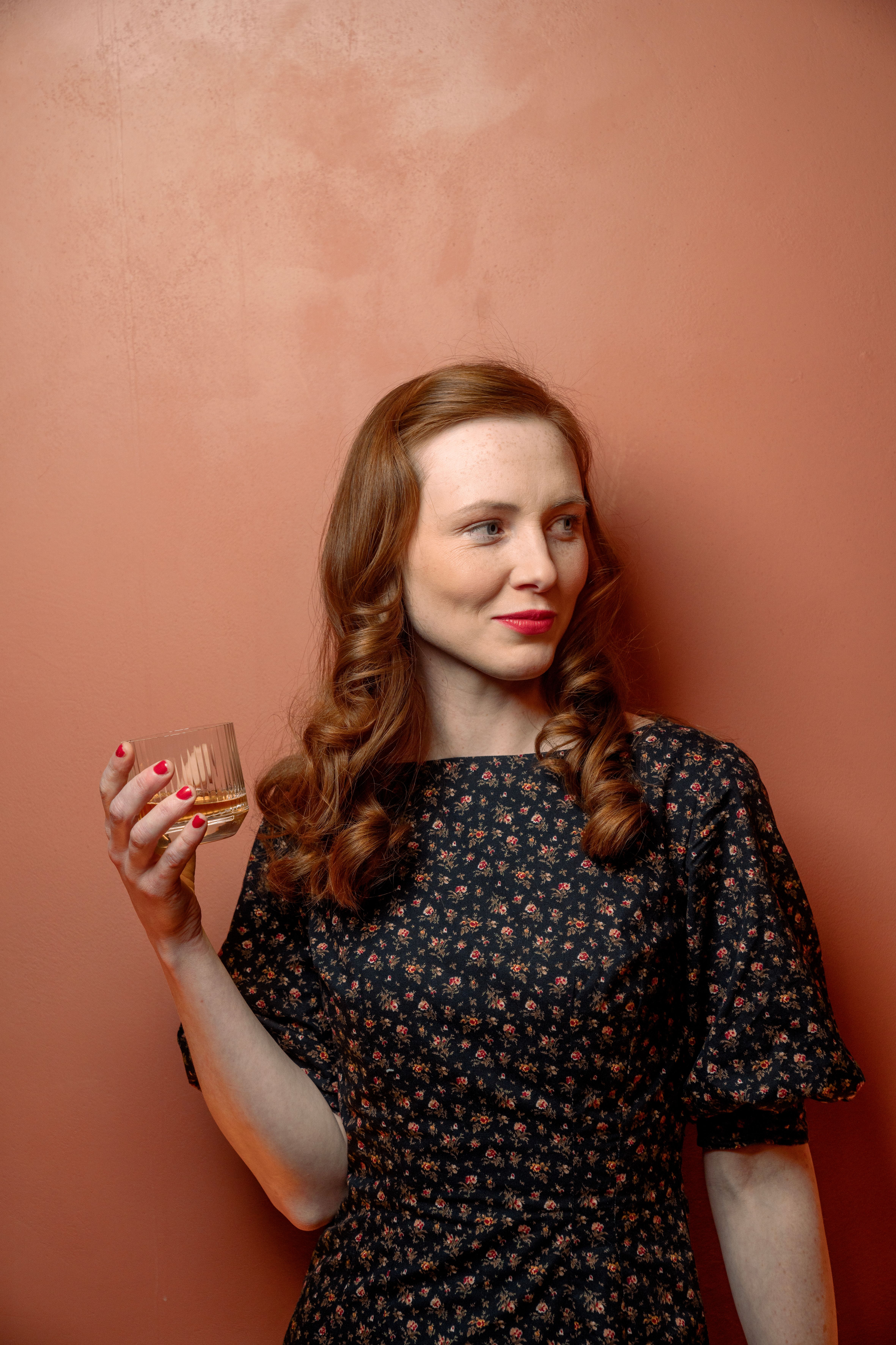 An elegant redhead woman poses with a glass against a warm background, exuding a vintage charm.