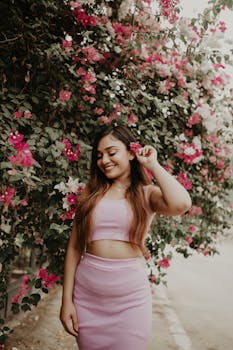 Portrait of a smiling woman in pink attire near a colorful flower bush outdoors.