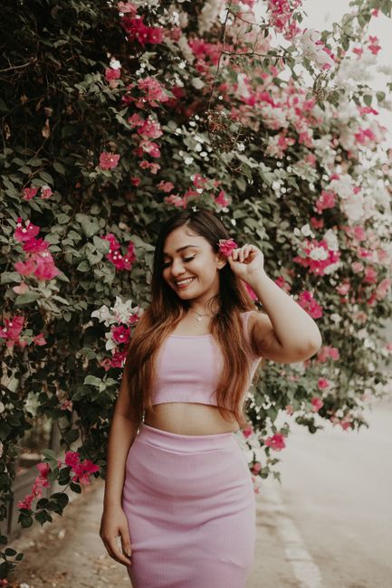 Portrait of a smiling woman in pink attire near a colorful flower bush outdoors.