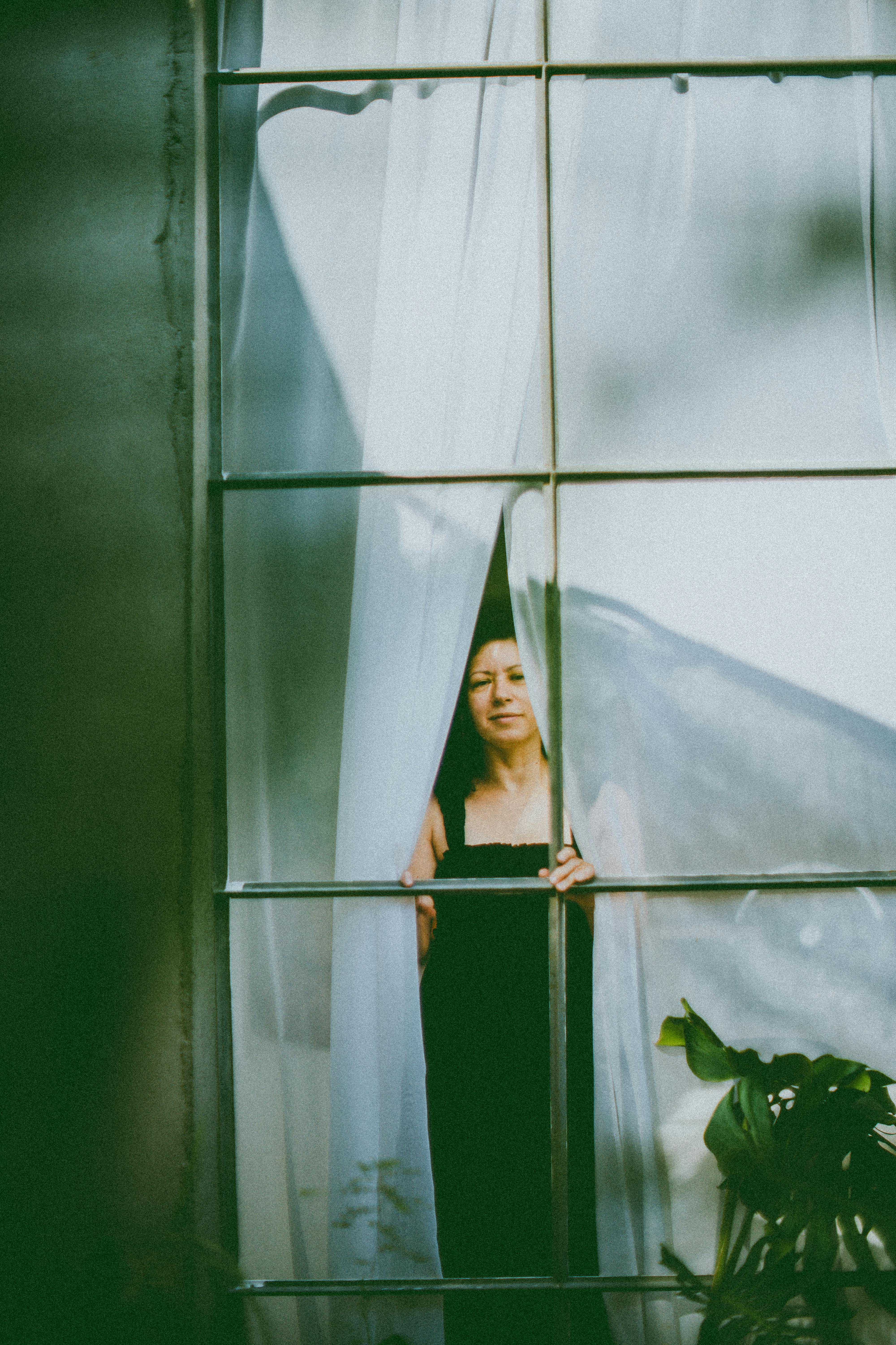 A woman peeks through sheer white curtains in an elegant setting.