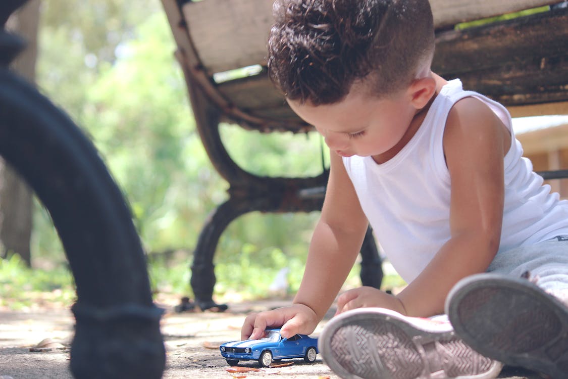 Мальчик в белой майке играет в синее купе под давлением возле коричневой деревянной скамьи днем