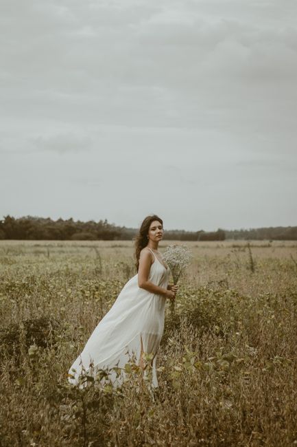 A bride in a wedding dress stands in a meadow under an overcast sky.