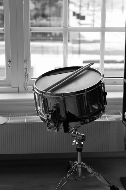 A monochrome image of a snare drum with drumsticks, placed by a window.