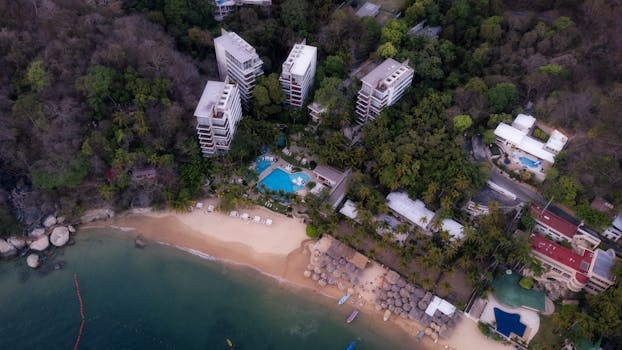 Drone shot of a beachfront resort with a pool in Acapulco, Mexico, surrounded by lush greenery and ocean views.