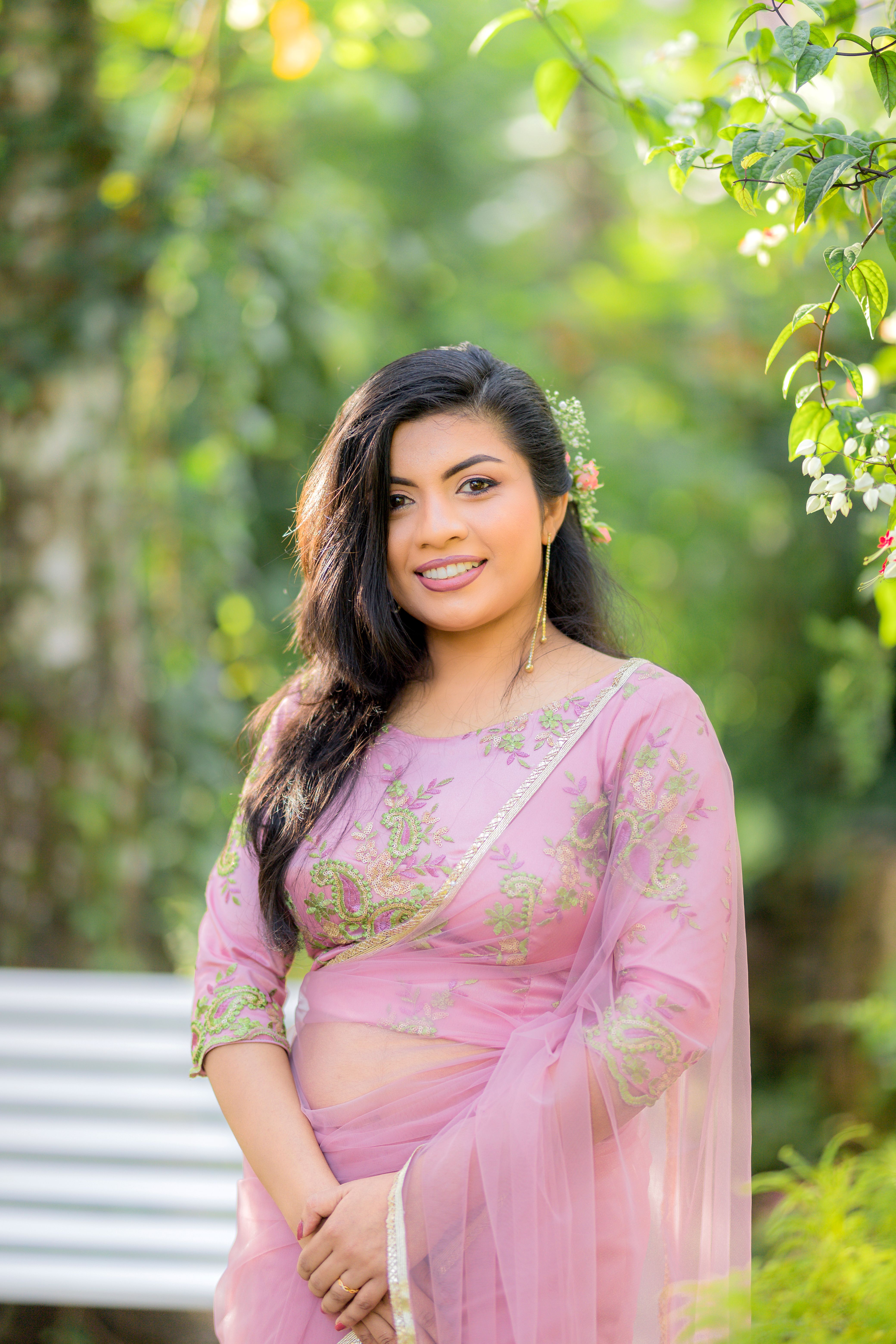 Woman in pink saree smiling in a sunlit garden setting, peaceful and elegant.