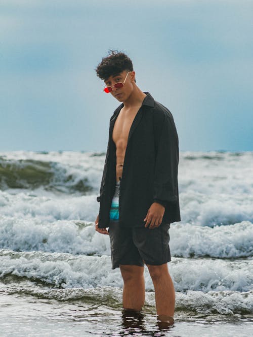 Young Man in a Shirt and Swimming Shorts Standing Ankles Deep in the Sea 