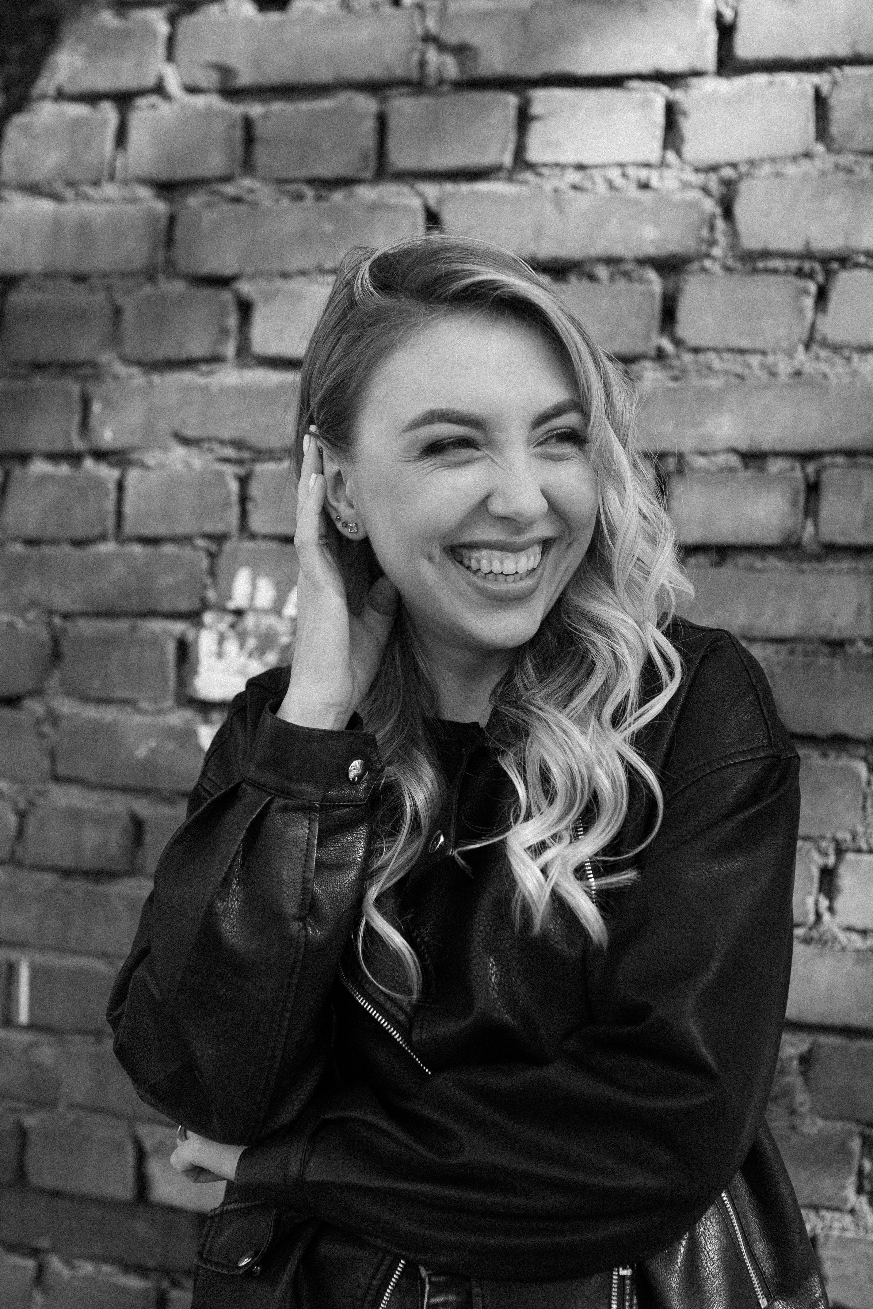 Elegant woman with long hair and leather jacket smiling by a brick wall in Russia.