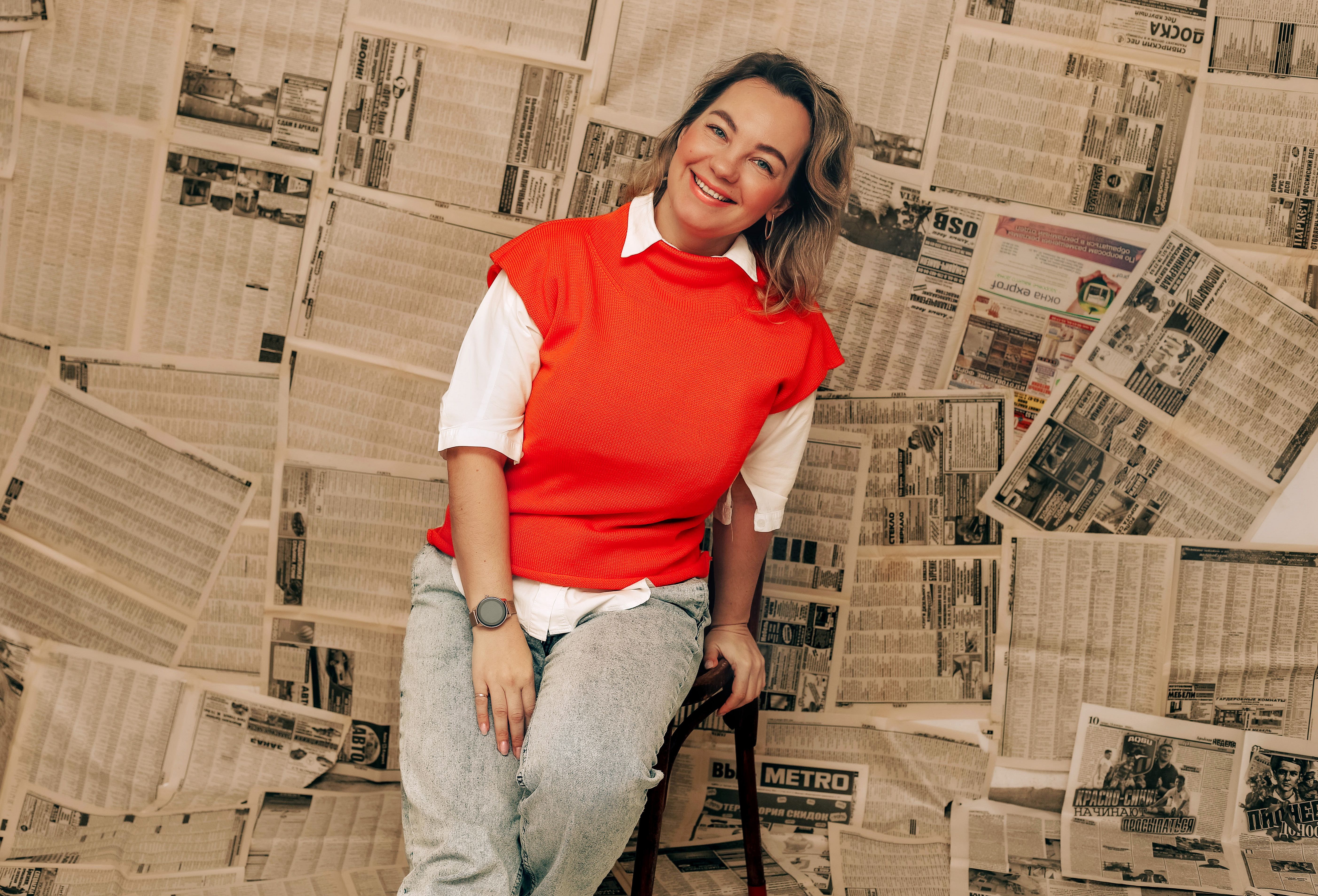 Fashionable woman with a joyful smile sitting against a wall covered in newspapers, creating a vintage vibe.