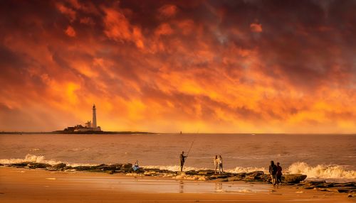 Free Fishermen by a lighthouse under a fiery sunset sky, creating a dramatic coastal scene. Stock Photo