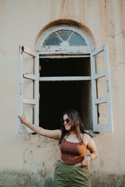 A young woman stands smiling by an open, rustic window, exuding charm.