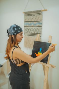 A young woman artist with a bandana paints on an easel in a cozy indoor setting, showcasing creativity.