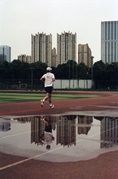 A man jogging on a track with city skyscrapers and a puddle reflection, capturing urban fitness