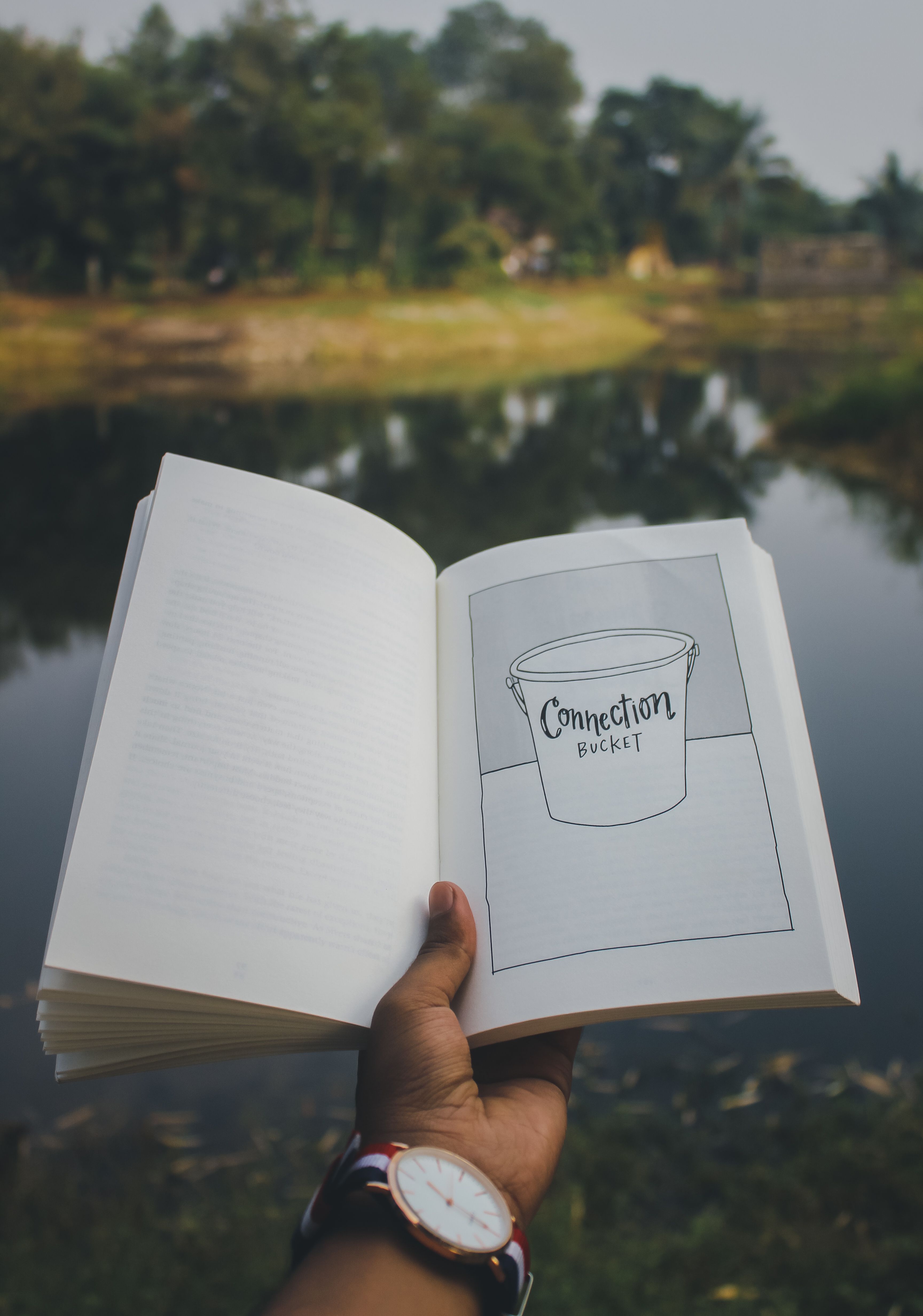 A man holds an open book by a serene lake, featuring a connection bucket sketch.