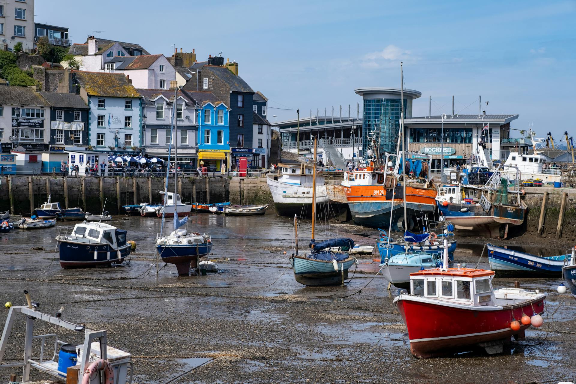 Vibrant coastal scene of Brixham Harbor at low tide, showing moored fishing boats and colorful waterfront buildings.
