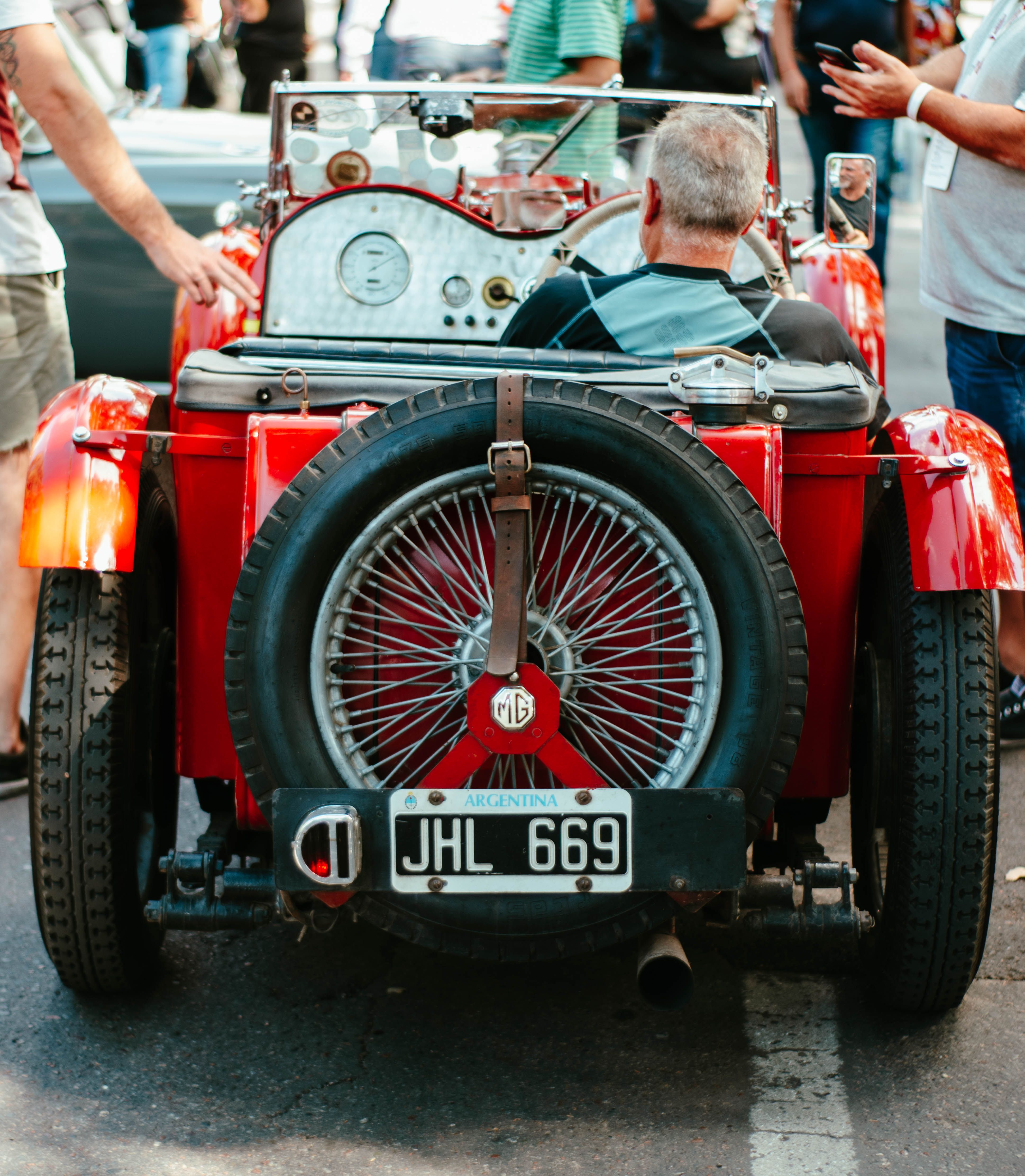 A man driving a classic red convertible car in a lively urban environment.