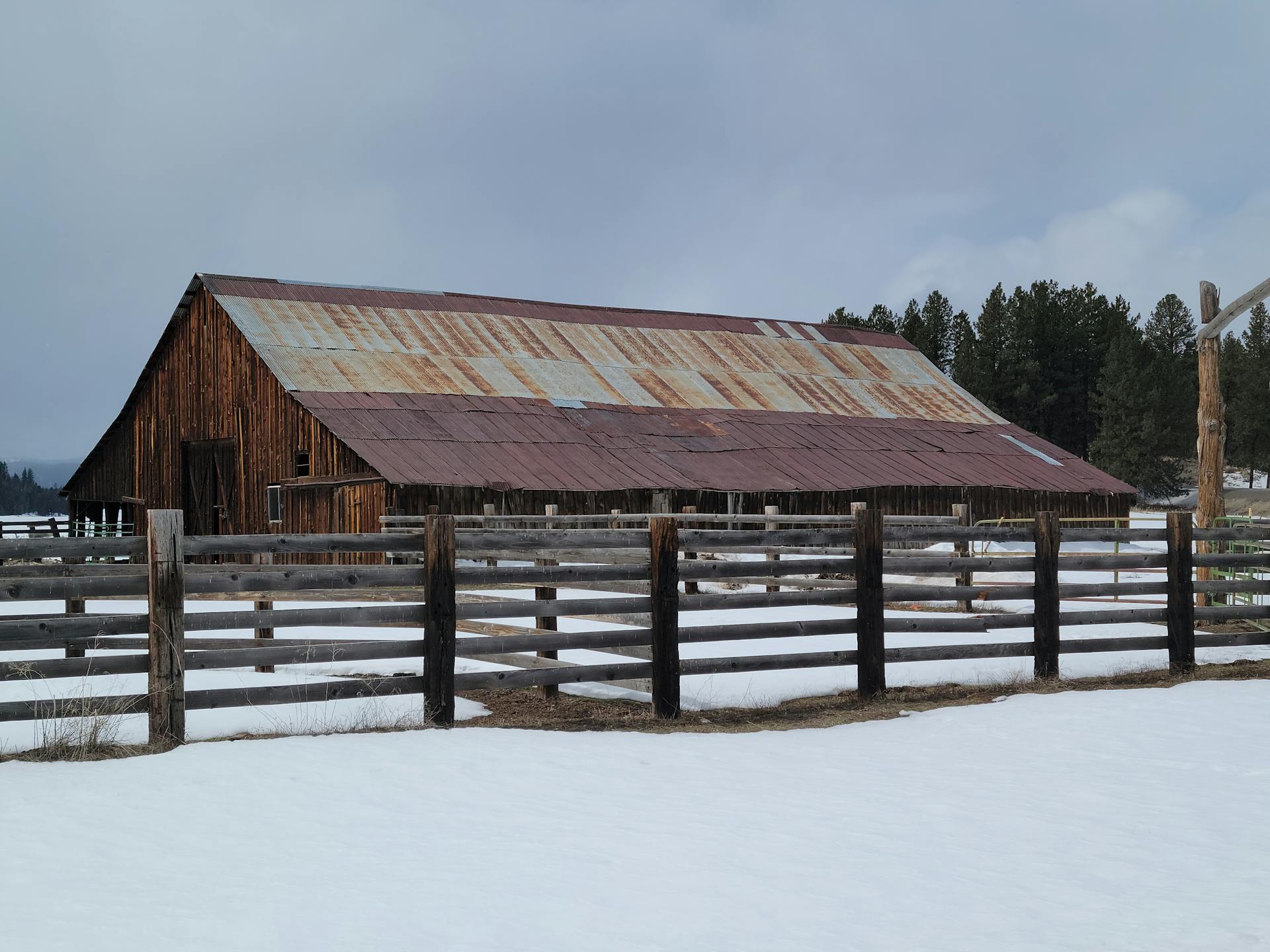 Wooden barn with rusted metal roof surrounded by snow and fence in a winter landscape.