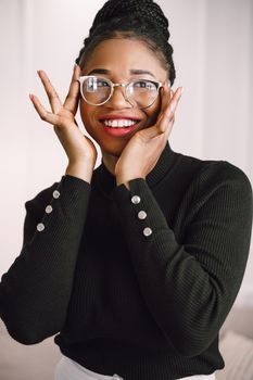 Joyful woman wearing glasses and sweater, smiling with hands on cheeks indoors.