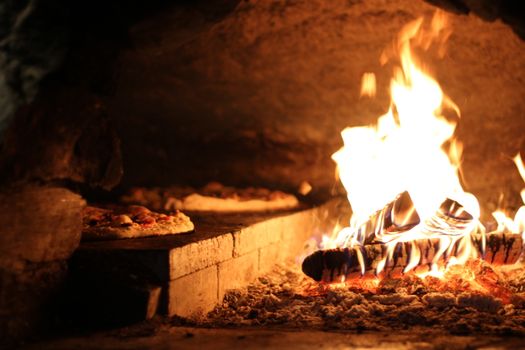 A close-up of a rustic wood-fired oven with pizzas near vibrant flames.