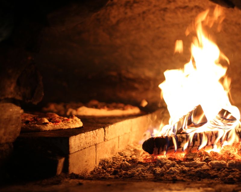 A close-up of a rustic wood-fired oven with pizzas near vibrant flames.