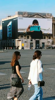 Two women walking by a large urban billboard in a city plaza.