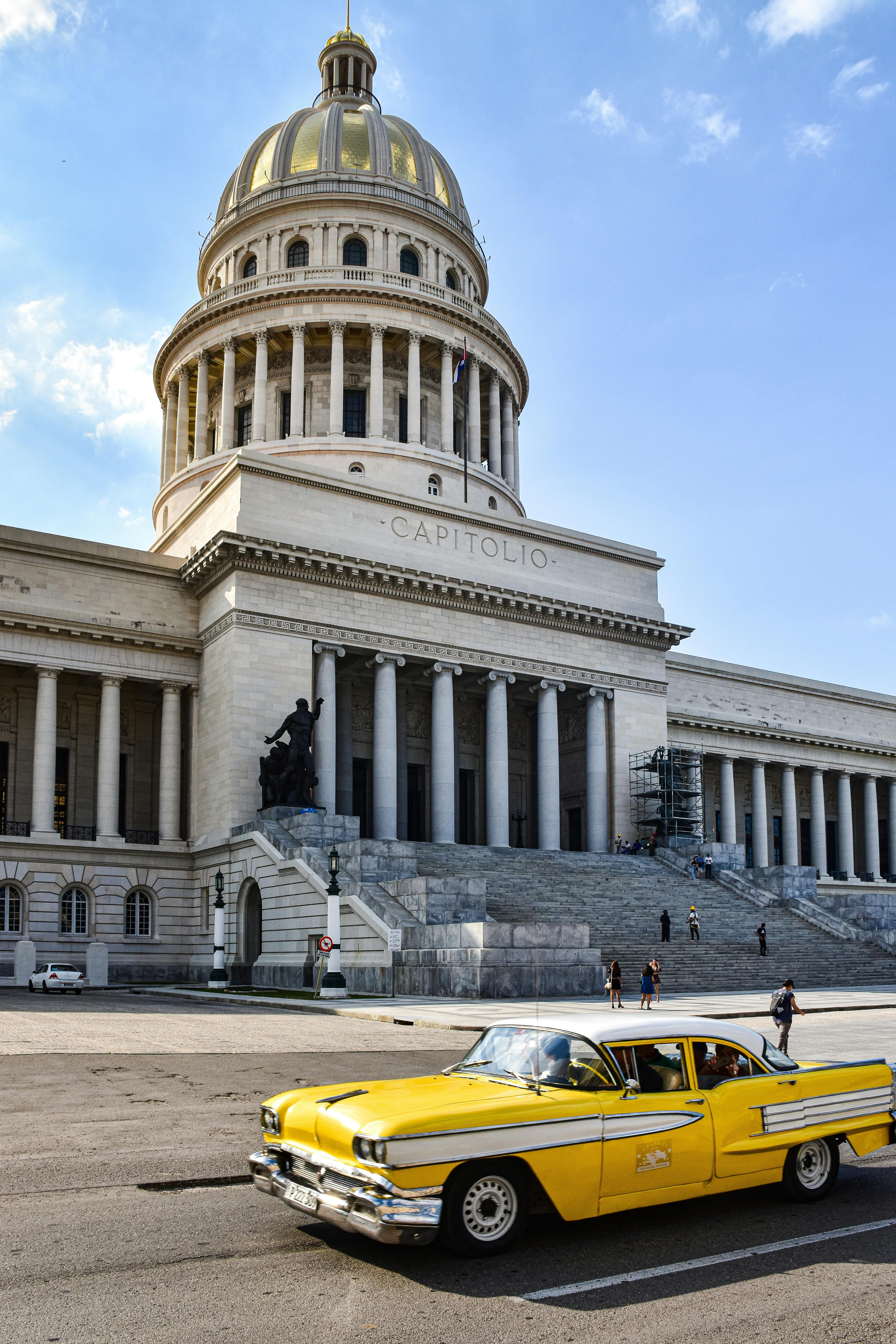 Classic yellow car driving past the El Capitolio in Havana, Cuba, featuring a clear blue sky.