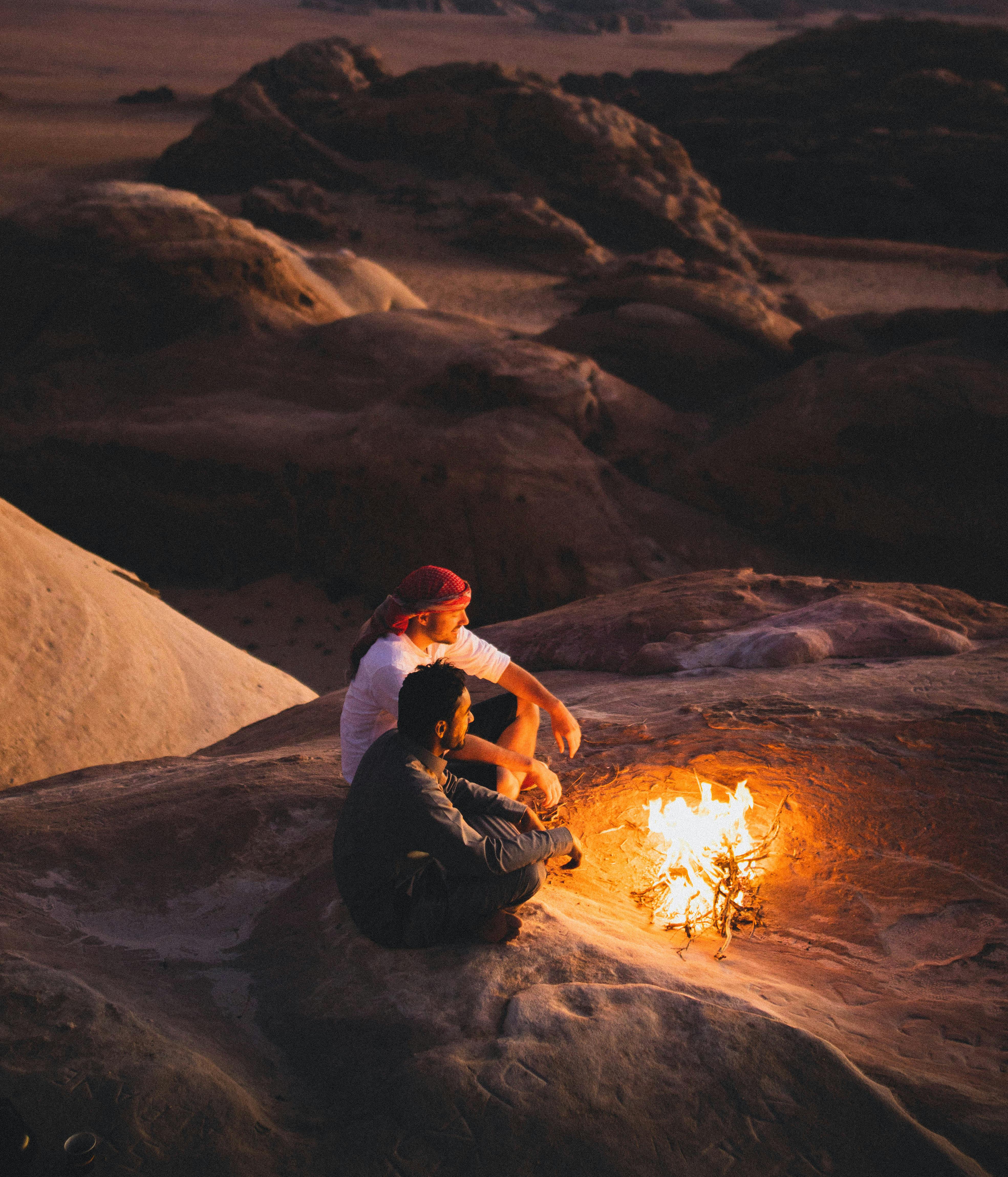 Free Two men sitting by a campfire against the stunning backdrop of Wadi Rum desert at sunset. Stock Photo