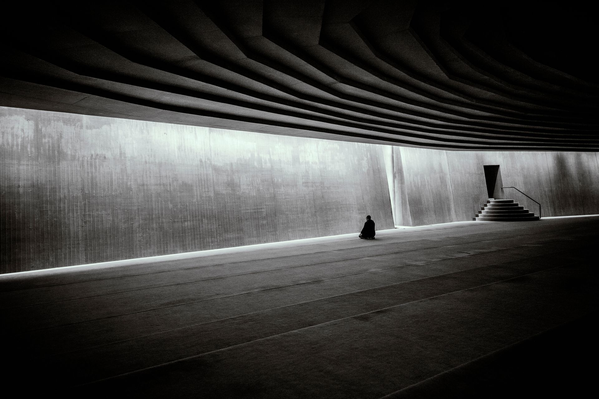 A lone person meditates in a vast, modern, minimalist indoor hall with dramatic architecture.