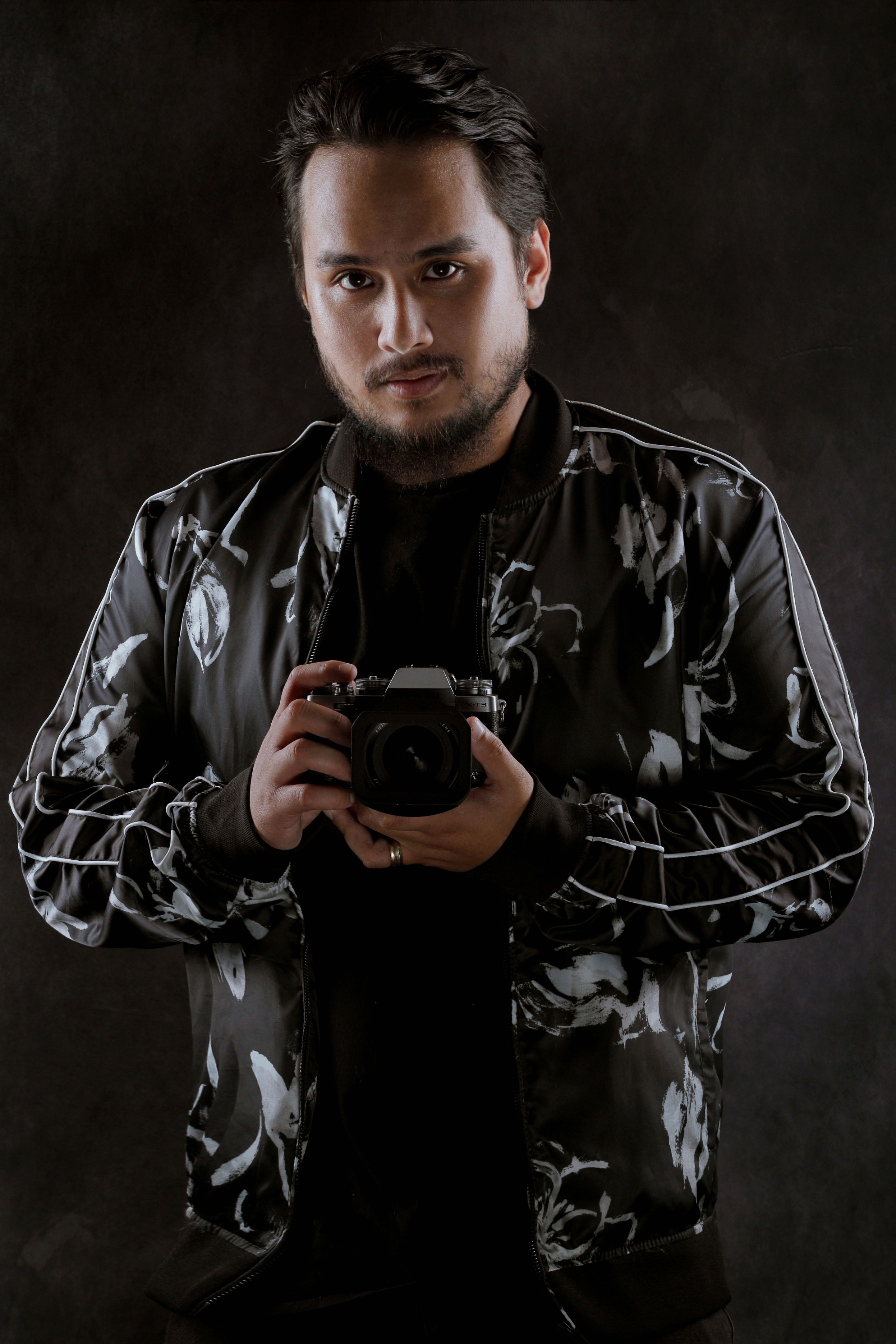 A photographer with a camera stands against a black background, exuding confidence and focus.