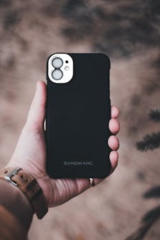A person's hand holding a smartphone in a Sandmarc case, outdoors.