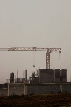 A misty, early morning view of a construction site with a crane and workers building amidst dense fog.