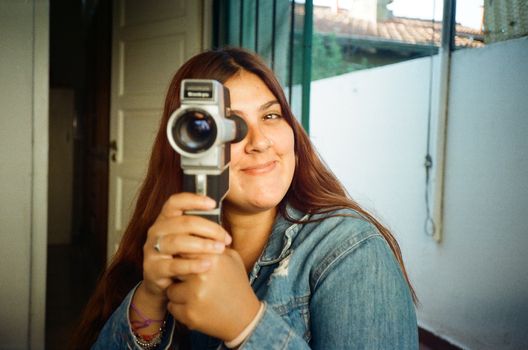 Smiling young woman in denim jacket using a vintage camcorder indoors in Mar del Plata.