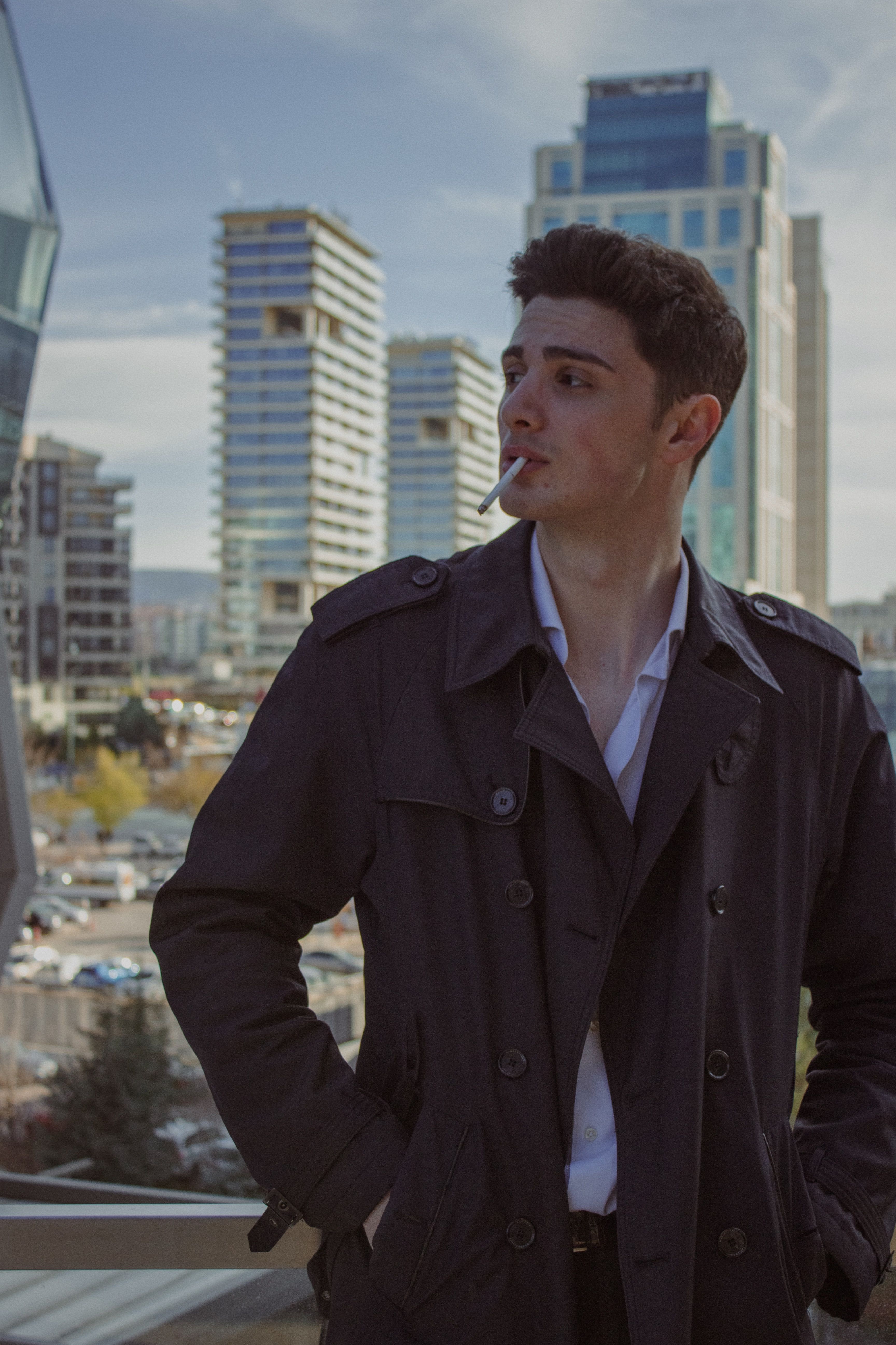 A man in a dark coat smokes with Ankara's modern skyline in the background.