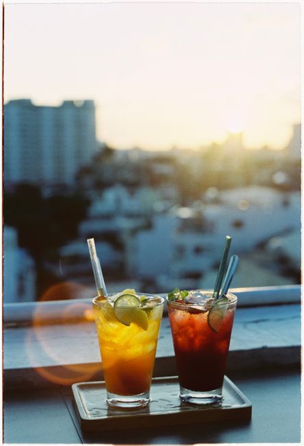 Two colorful cocktails with lime garnish on a rooftop during sunset, capturing an urban skyline in the background.