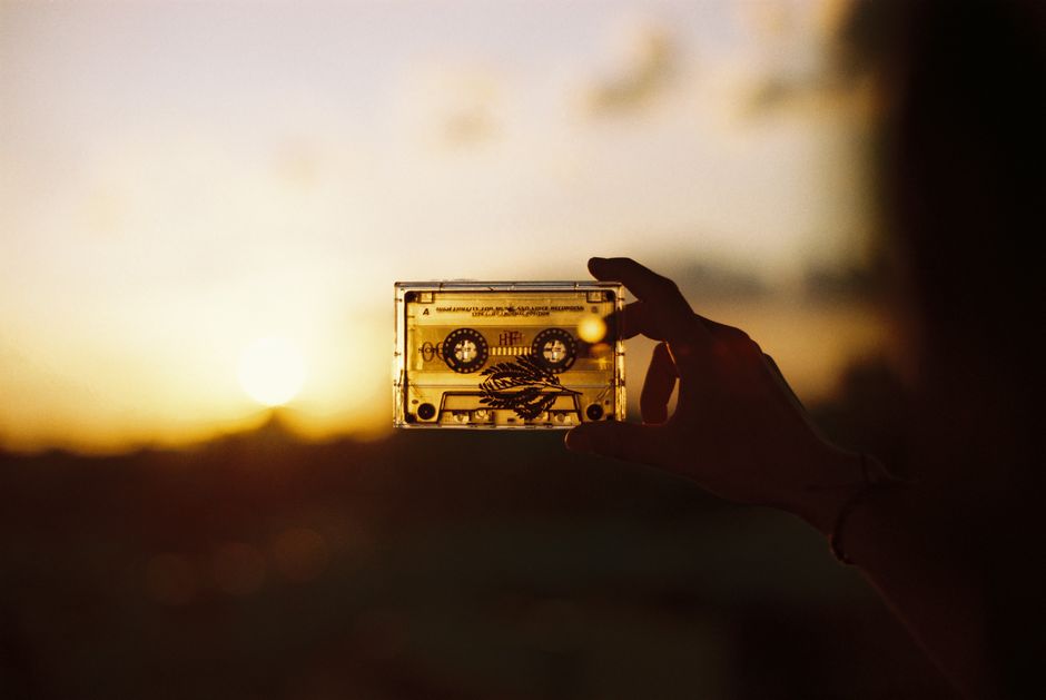 Person holding a cassette tape against a warm, sunset backdrop, creating a nostalgic silhouette effect.