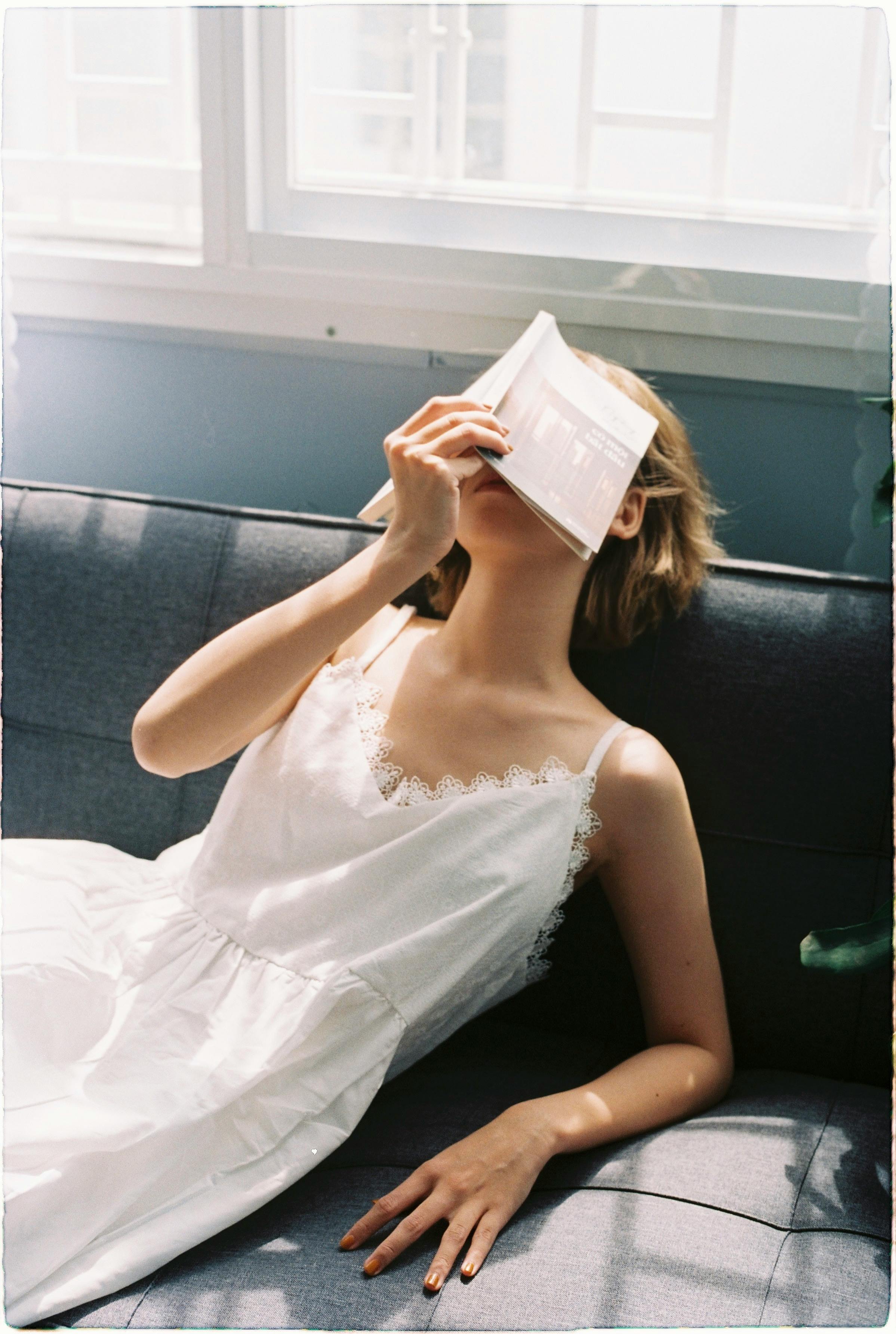 A woman in a white dress rests on a sofa, holding a book on her face, enjoying a sunny day indoors.