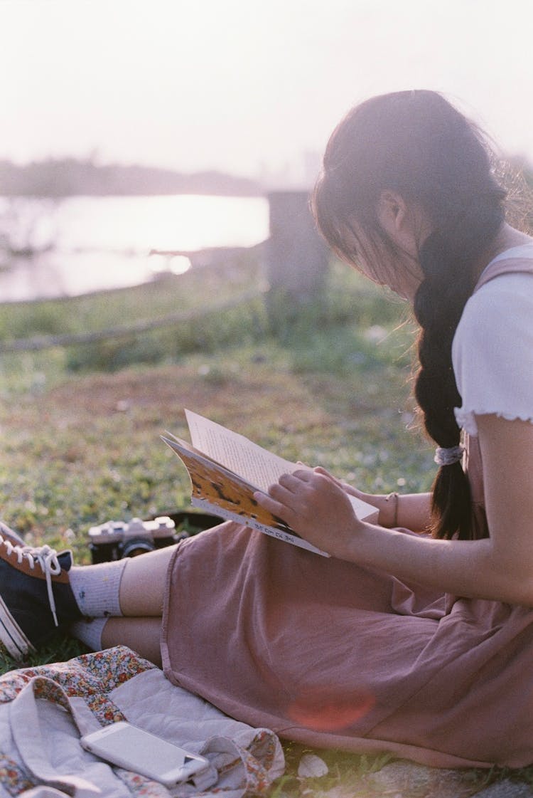 Girl Sitting On Grass Near River Reading Book