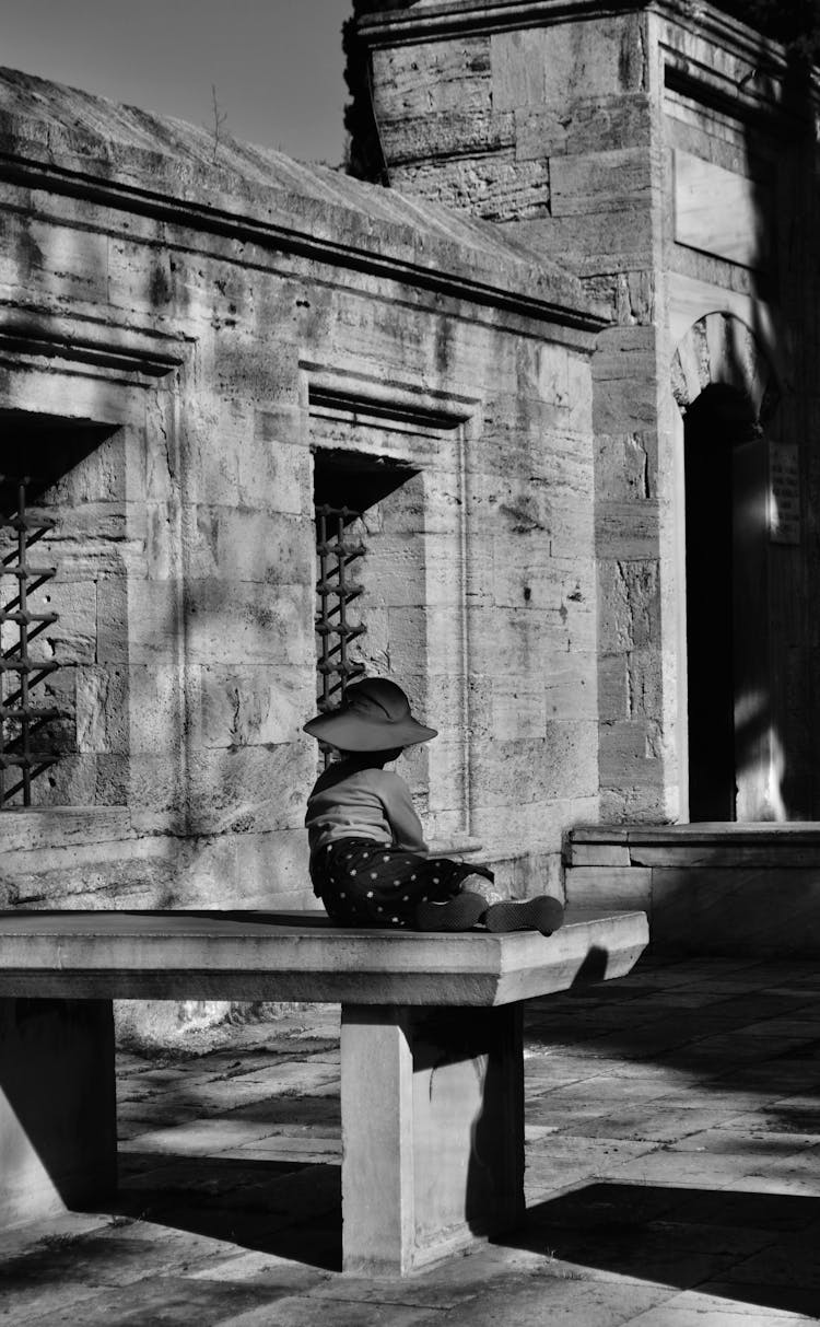 Child Sitting On Stone Table Near Building In Black And White