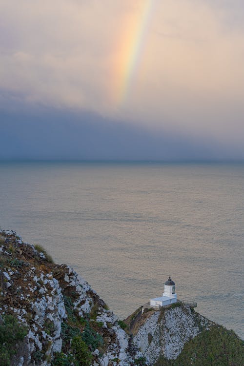 Rainbow over Clouds over Sea Shore with Lighthouse on Hill with Rocks