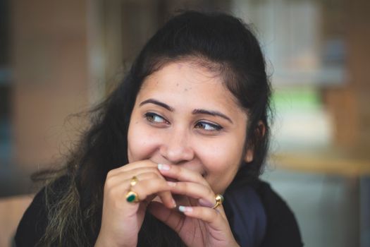 A candid portrait of a smiling young woman covering her mouth in a thoughtful pose indoors.