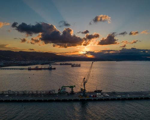 Free A breathtaking view of cranes and ships at a harbor during sunset, reflecting on the calm sea. Stock Photo