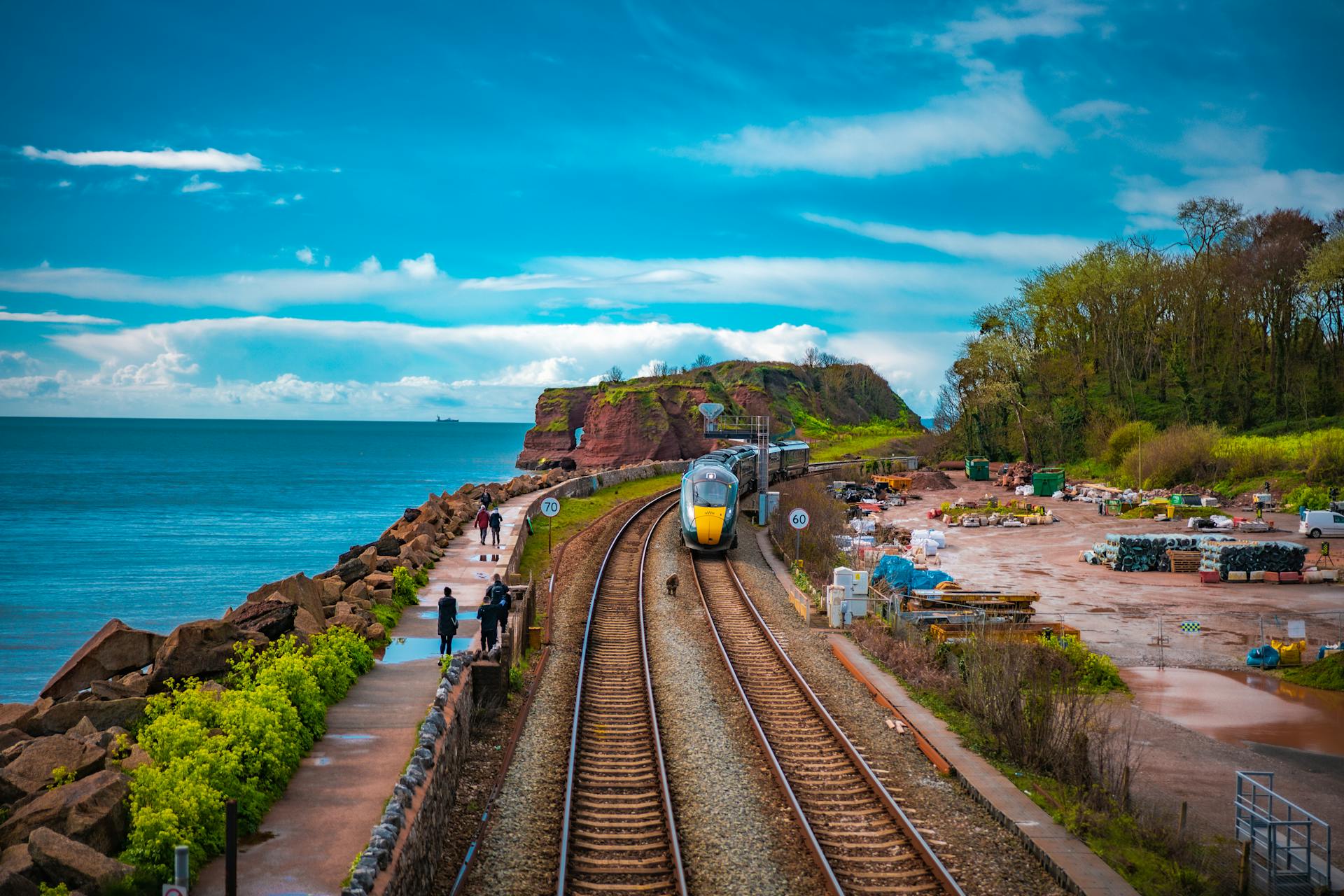 A scenic coastal railway with a modern train, adjacent to a walkway and sea view, showcasing nature and transportation.