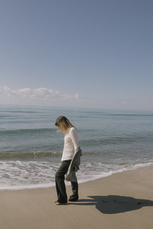 Free A woman in stylish attire walking along a sunny beach shore, capturing a serene moment. Stock Photo