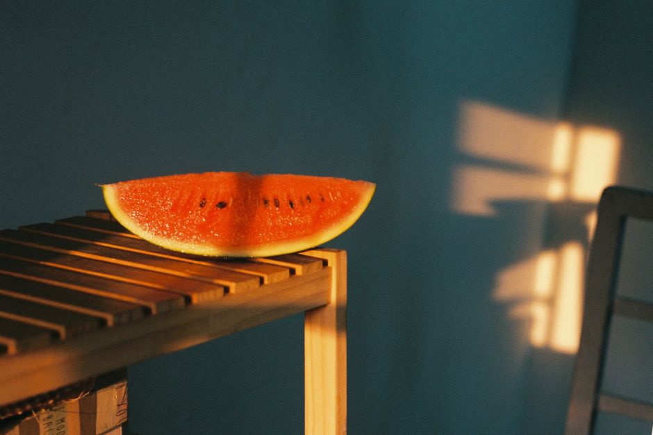 A slice of watermelon rests on a wooden table with dramatic sunlight casting shadows indoors.
