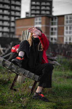 一位身穿红色夹克的年轻女子，在城市公园的长凳上用手遮住脸摆姿势