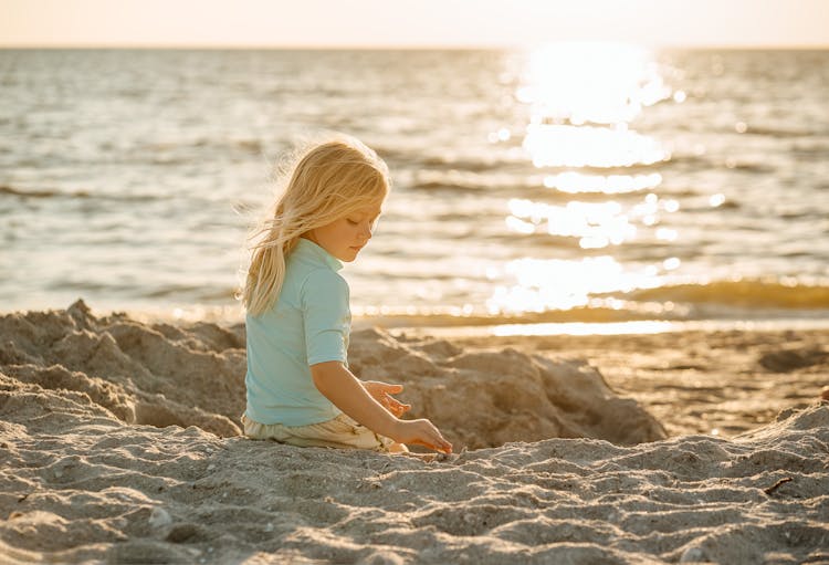 Blond Girl Sitting On A Sandy Beach, And Sun Reflecting In The Sea