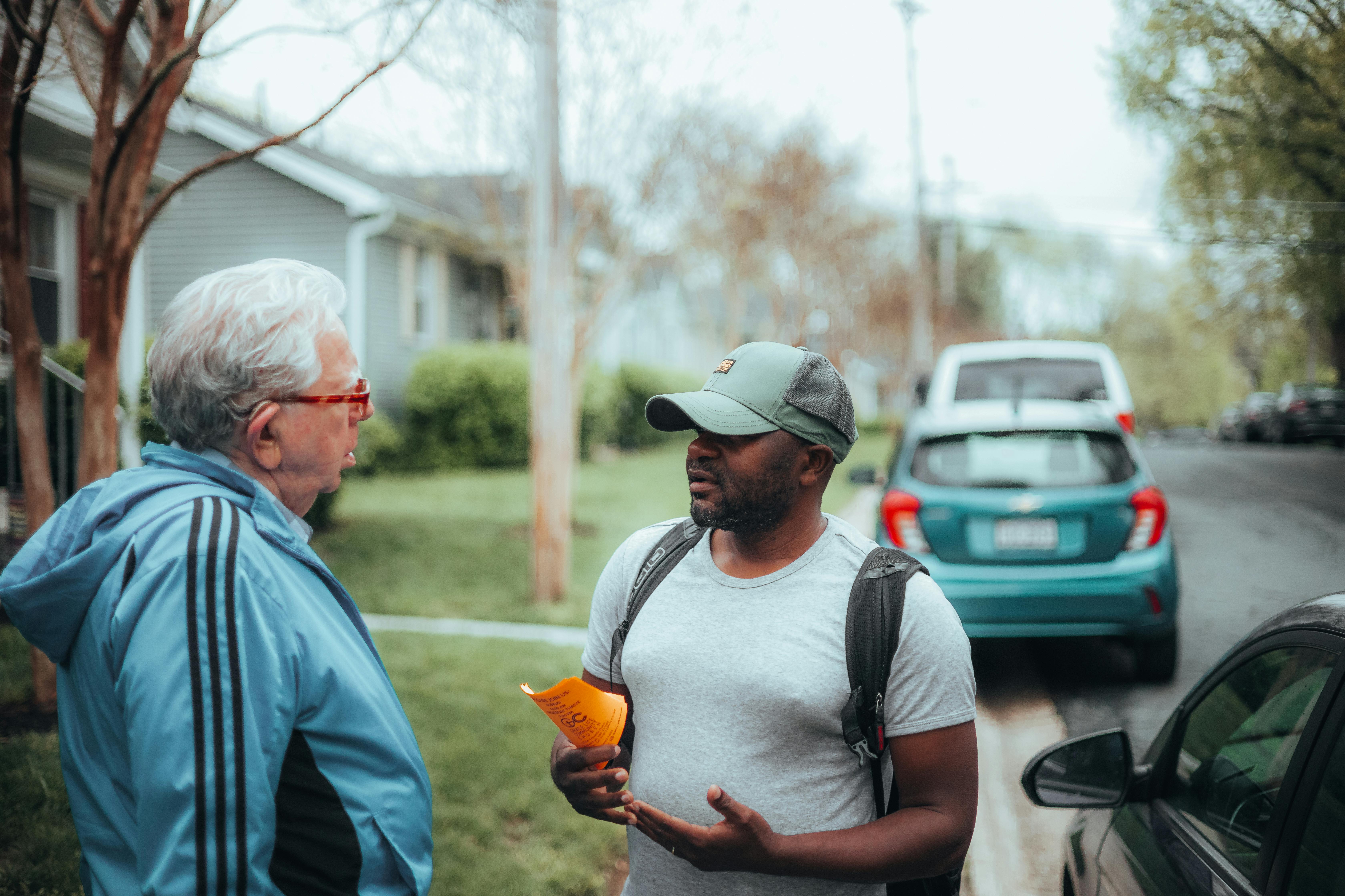 Two men engaged in conversation on a suburban sidewalk with parked cars.