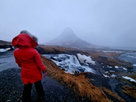 A woman in a red coat stands in front of a waterfall
