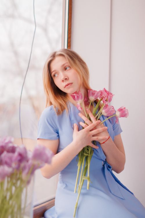 Free A thoughtful woman in blue holding pink tulips by a window indoors. Stock Photo