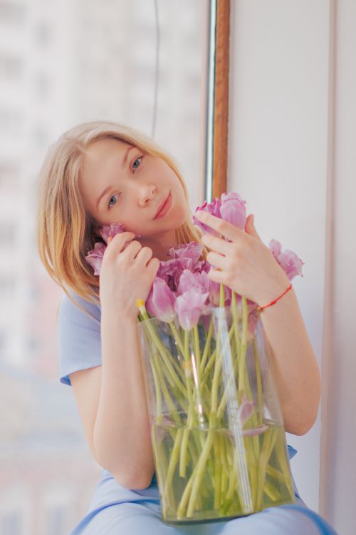 Free Blonde woman in blue dress holding pink tulips by window indoors. Stock Photo